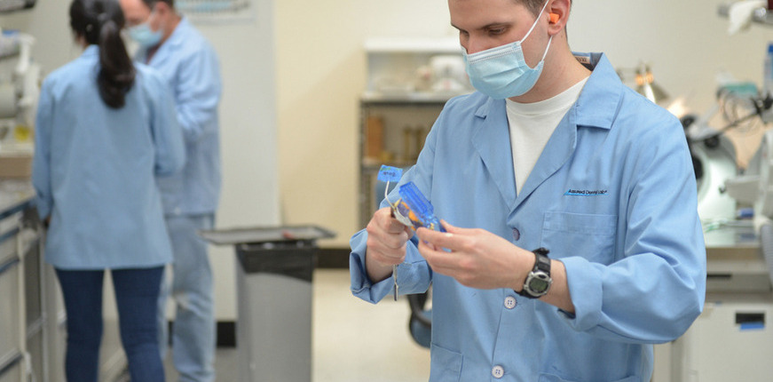 A dental technician in a light blue Assured Dental Lab coat and mask holds a blue object, possibly dental prosthetic or restoration made in the lab.