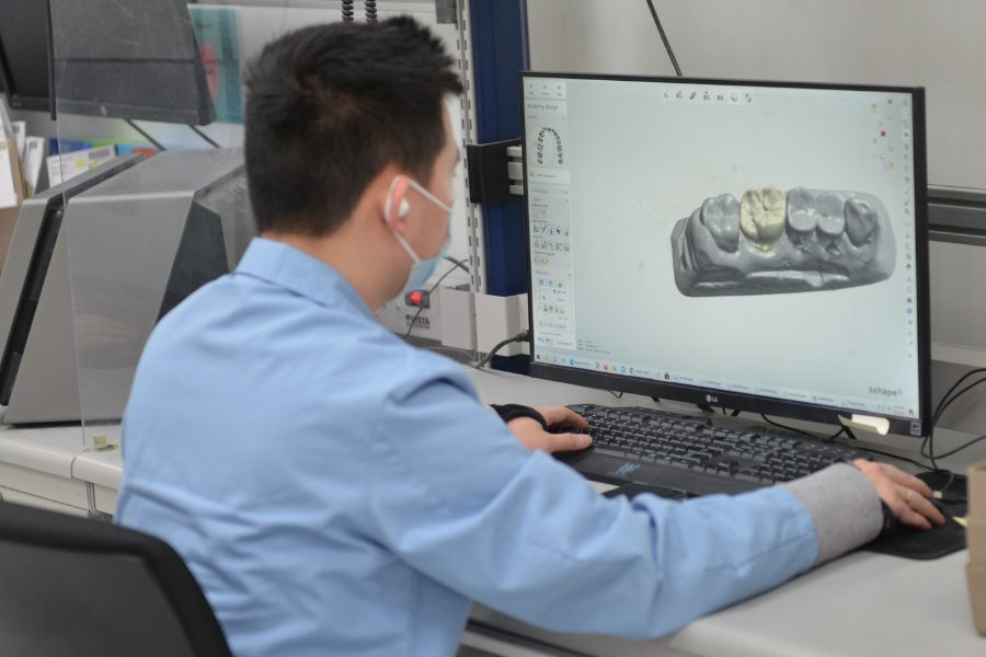 A dental technician in a blue shirt sits in front of a computer, wearing a mask and examining a dental restoration model.