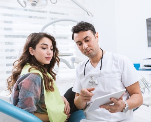 A dentist is pointing to a clipboard, as if showing x-rays or steps to a procedure. A patient sits on a dental chair looking at the clip board intently.