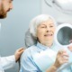 An elderly woman is holding a mirror and smiling, observing her new zirconia dental implants as a dentist watches close by.