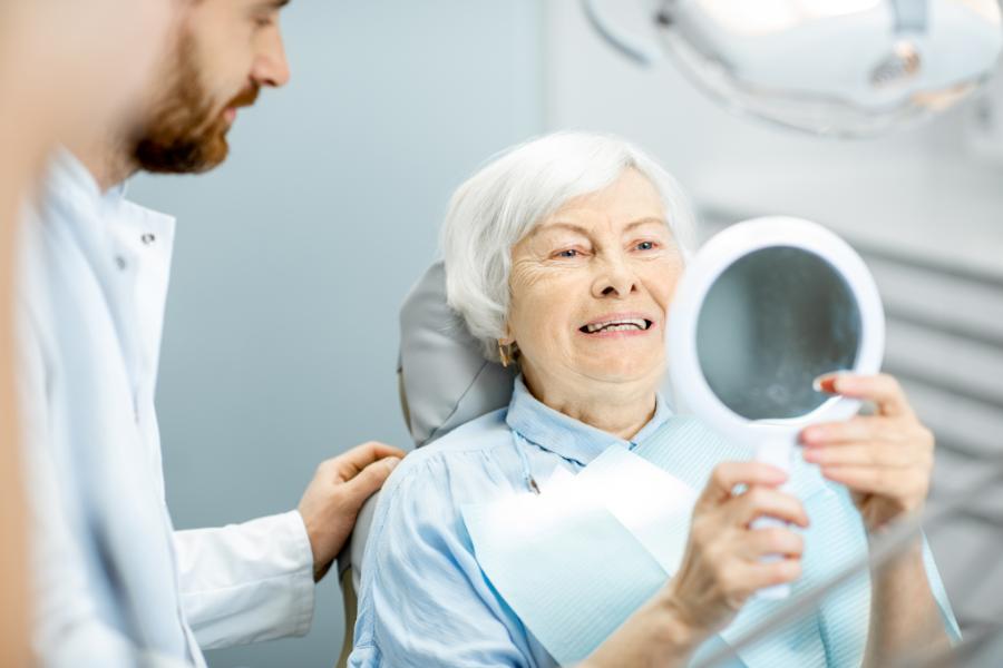 An elderly woman is holding a mirror and smiling, observing her new zirconia dental implants as a dentist watches close by.