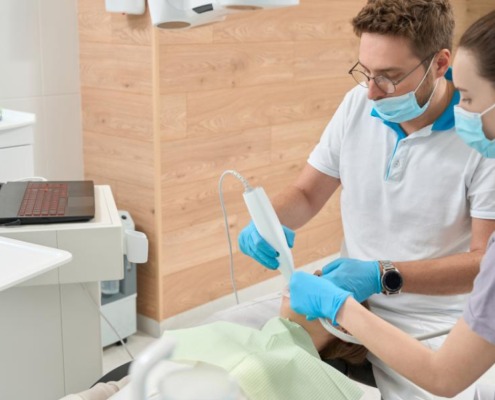 A man holds a piece of dental equipment and is examining a patient's teeth, with a dental assistant watching closely.