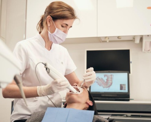 Professional dentist in mask and uniform using 3D scanner for digital impressions of patient and controlling process on laptop in modern clinic.