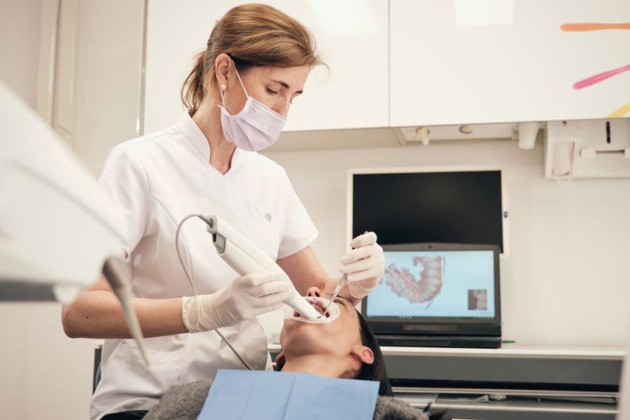 Professional dentist in mask and uniform using 3D scanner for digital impressions of patient and controlling process on laptop in modern clinic.